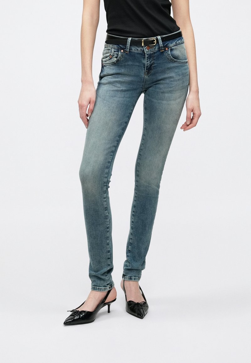 Femme portant un jean skinny bleu délavé avec une ceinture noire et des escarpins noirs à bouts pointus, debout devant un fond uni.