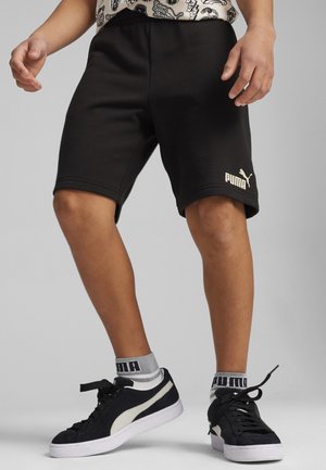 Pantalones cortos deportivos de algodón negros con un ajuste ajustado, que presentan un logo dorado de PUMA. Zapatillas negras y blancas combinadas con calcetines acanalados grises y negros.