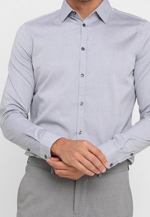 Hombre que lleva una camisa de vestir gris claro abotonada y pantalones grises a juego, ajustándose el puño izquierdo con ambas manos.