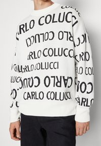 Suéter blanco con el texto negro "CARLO COLUCCI" repetido en diferentes orientaciones. Textura suave, cuello redondo y mangas largas.