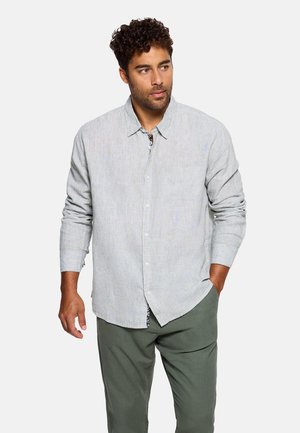 Hombre con cabello rizado que lleva una camisa de manga larga gris claro abotonada y pantalones verde oliva, mirando hacia un lado, con una mano en el bolsillo.