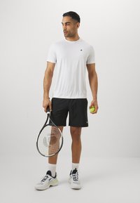 Weißes kurzärmliges Sportshirt, schwarze Shorts, hält ein Tennisracket und einen Ball. Die Schuhe haben weiße, graue und marineblaue Akzente.
