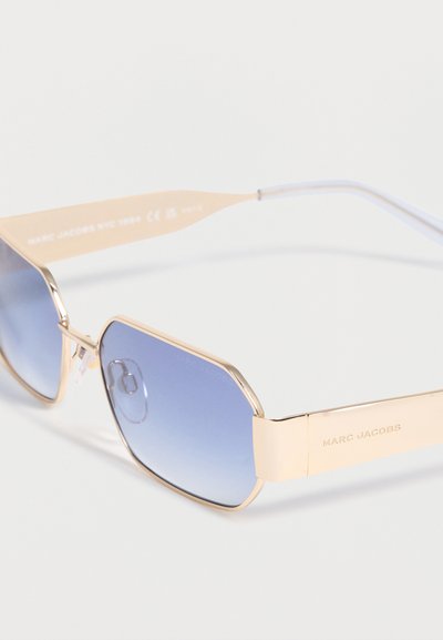 Γυαλιά ηλίου με χρυσό σκελετό και φακούς με μπλε διαβάθμιση, με χαραγμένο λογότυπο "Marc Jacobs" στα πλαινά, παρουσιάζονται μπροστά σε λευκό φόντο.