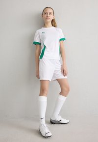 Valge sportlik komplekt, mis sisaldab lühikeste varrukatega tippi roheliste detailidega ja sobivaid lühikesi pükse. Välimuse täiendavad valged sokid ning musta ja valge triibuga jalgpalliketsid.