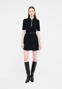 Abito polo nero con maniche corte, zip frontale e una cintura aderente. Abbinato a stivali neri lucidi fino al ginocchio, creando una silhouette elegante.