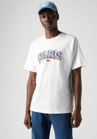 Camiseta blanca de manga corta con un gráfico multicolor de "PARIS", combinada con unos vaqueros de denim azul y una gorra azul clara.