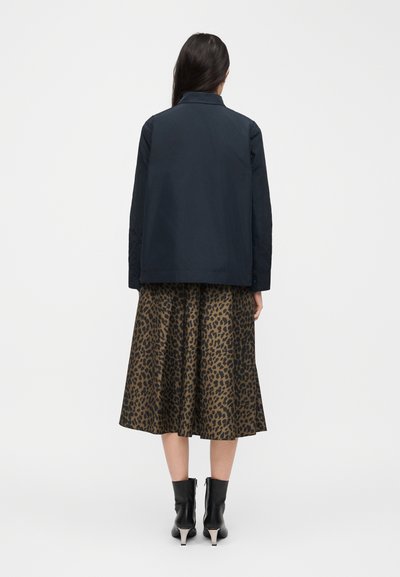 Γυναίκα που στέκεται με την πλάτη στον θεατή, φορώντας σκούρο μπουφάν, μίντι φούστα με λεοπάρ μοτίβο και μαύρα μποτάκια με τακούνια.