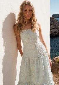 Jeune femme aux longs cheveux ondulés portant une robe d'été bustier brodée, debout contre un mur blanc près d'une côte rocheuse et de la mer.