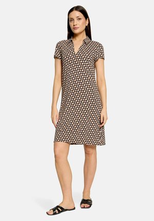 Femme debout portant une robe à motif à manches courtes, longueur genou, avec un col et des sandales noires à enfiler sur un fond uni.
