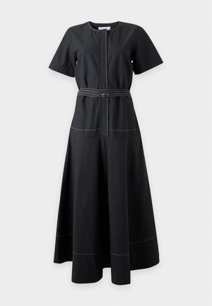 Femme aux longs cheveux foncés portant une robe midi noire ceinturée, tenant un sac à main marron et des sandales noires à bout ouvert sur un fond uni.