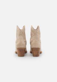 Béžové semišové kotníkové boty s naskládaným dřevěným podpatkem. Mají špičatou špičku a ozdobné švy na bocích. Měkká textura s hladkým povrchem.