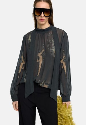 Kobieta w dużych czarnych okularach przeciwsłonecznych, przezroczystej czarnej bluzce z abstrakcyjnymi beżowymi wzorami, czarnych spodniach, trzymająca żółtą puszystą torebkę.