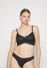 Svart spiss bralette med bølgete kant og justerbare stropper, parret med sømløse svarte bikinitruser. Myk, teksturert stoff.