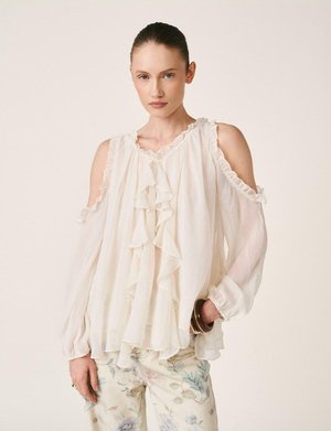 Mujer con blusa blanca de hombros descubiertos con volantes y mangas largas, y pantalones con estampado floral, de pie con una mano en el bolsillo.