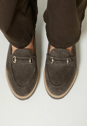 Pieds portant des mocassins en suède marron foncé avec un détail en métal, assortis à un pantalon marron foncé visible d’en haut.