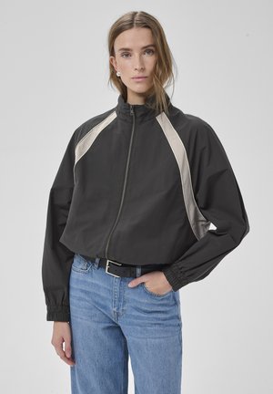 Femeie purtând o jachetă neagră cu fermoar și dungi bej pe umeri și blugi albaștri deschiși, stând cu o mână în buzunar, pe un fundal simplu.