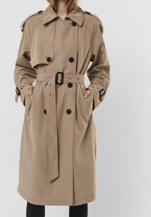 Kvinde iført en beige to-radet trenchcoat med sorte knapper og bælte i taljen, hænderne i lommerne, mod en ensartet lys baggrund.