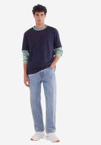 Marinblå t-shirt lager över en ljusgrön randig långärmad tröja; kombinerad med ljusblå jeans och vita sneakers med bruna sulor.