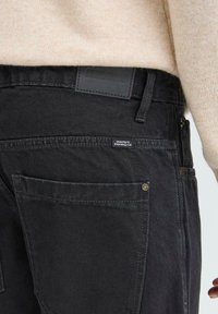 Pantaloni din denim negri cu o croială lejeră, cu un patch din piele, cusături vizibile și două buzunare pe spate cu accente din butoane de alamă.