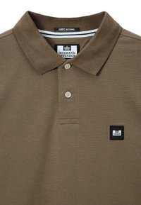 Bruine poloshirt van een gestructureerde stof, met twee knopen en een klein zwart logo op de patch. Heeft een ribbelkraag met witte accenten.