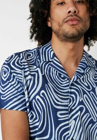 Kurzärmliges Hemd aus blauem Stoff mit weißen, wavy Linienmustern. Button-Down-Kragen und sichtbare Knöpfe auf der Vorderseite.