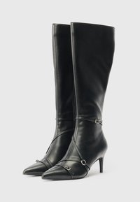 Botas de cuero negro hasta la rodilla con puntas afiladas, tacones delgados y correas decorativas con pequeñas hebillas metálicas sobre un fondo blanco.