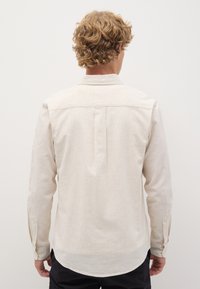Camisa de manga larga color beige claro, hecha de lino, con cuello estándar, puños abotonados y un pliegue central en la parte posterior para mayor comodidad.