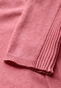 Dettaglio di un maglione rosa lavorato a maglia che mostra una texture liscia, bordi a coste sui polsini e sottili variazioni di colore in tutto il tessuto.