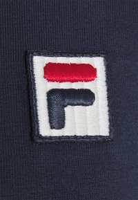 Stickad logotyp på en marinblå tyg med en röd och vit rektangulär överdel som har ett stiliserat bokstav 'F' i marinblått. Slät textur.