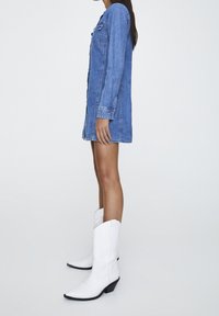 Robe chemise en denim bleu clair avec manches longues, poches avant et coupe droite. Associée à des bottes de cowboy blanches à bouts pointus.