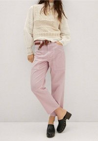 Pantalones de pierna ancha de color rosa claro con bolsillos laterales, un suéter de punto texturizado en color crema y zuecos negros con tacón de madera y tachuelas doradas.