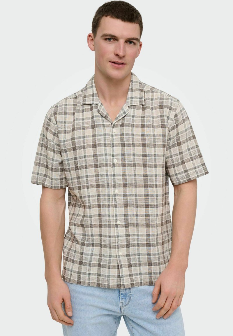 Mand iført kortærmet beige, brun og cremefarvet ternet skjorte med knapper og lyseblå jeans, stående mod en ensfarvet baggrund.