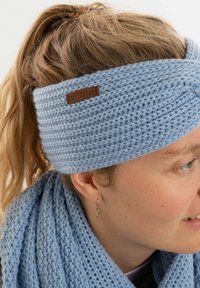 Donna con capelli castano chiaro che indossa una fascia per capelli lavorata a maglia di colore azzurro chiaro e una sciarpa abbinata, con un piccolo orecchino a forma di foglia visibile.