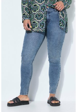 Frau trägt blaue enge Jeans, langärmliges Hemd mit Blumenmuster, schwarze Pantoletten und gelben Nagellack an den Zehen und Fingern.