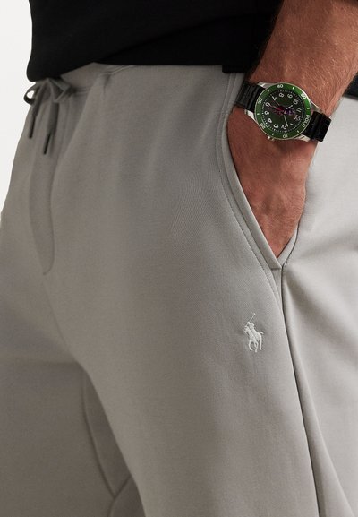 Γκρι παντελόνια τζόγκινγκ με λεία υφή, ρυθμιζόμενη μέση με κορδόνι και κεντητό λογότυπο. Φοριούνται με ένα μαύρο ρολόι που έχει πράσινο καντράν.