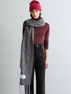 Frau trägt eine rote Strickmütze, ein bordeauxrotes Langarmshirt, schwarze hoch taillierte Hose und einen langen grauen Schal mit weißem Etikett.