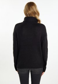 Schwarzer, grob gestrickter Pullover mit hohem Kragen, langen Ärmeln und geripptem Saum. Über dunklen Jeans getragen, von hinten angezeigt.