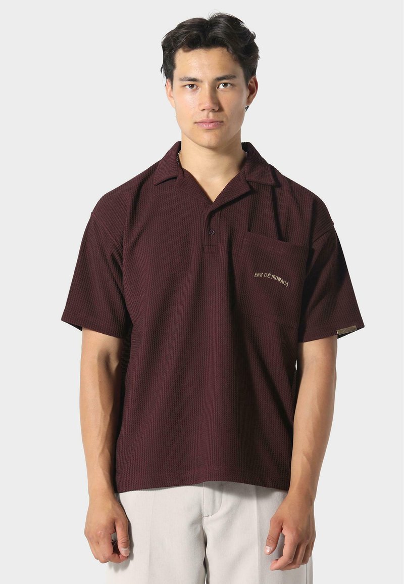 Polo shirt corto color marrone bordò con un motivo a trama, dotato di taschino sul petto e dettagli ricamati. Abbinato a pantaloni di colore chiaro.