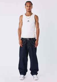 Camiseta sin mangas blanca con logo negro, vaqueros anchos azul oscuro y zapatillas deportivas blancas. Detalles destacables incluyen costuras visibles en los vaqueros.