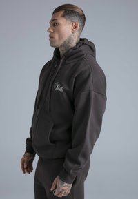 Mörkbrun hoodie av mjukt tyg med framficka, dragsko och broderad logotyp på bröstet.