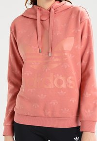 Rosa hoodie med en stor Adidas-logotyp, tillverkad av mjukt tyg med ett subtilt mönster av små logotyper i hela plagget. Justerbar dragsko i huvan.