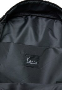 Intérieur du sac à dos noir en tissu nylon lisse. Comprend une étiquette avec un design signature. Fermeture éclair le long du bord supérieur.