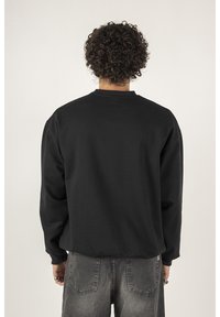 Felpa nera con collo rotondo, maniche lunghe e vestibilità rilassata. Il tessuto appare morbido con una texture liscia, indossata con jeans in denim grigi.
