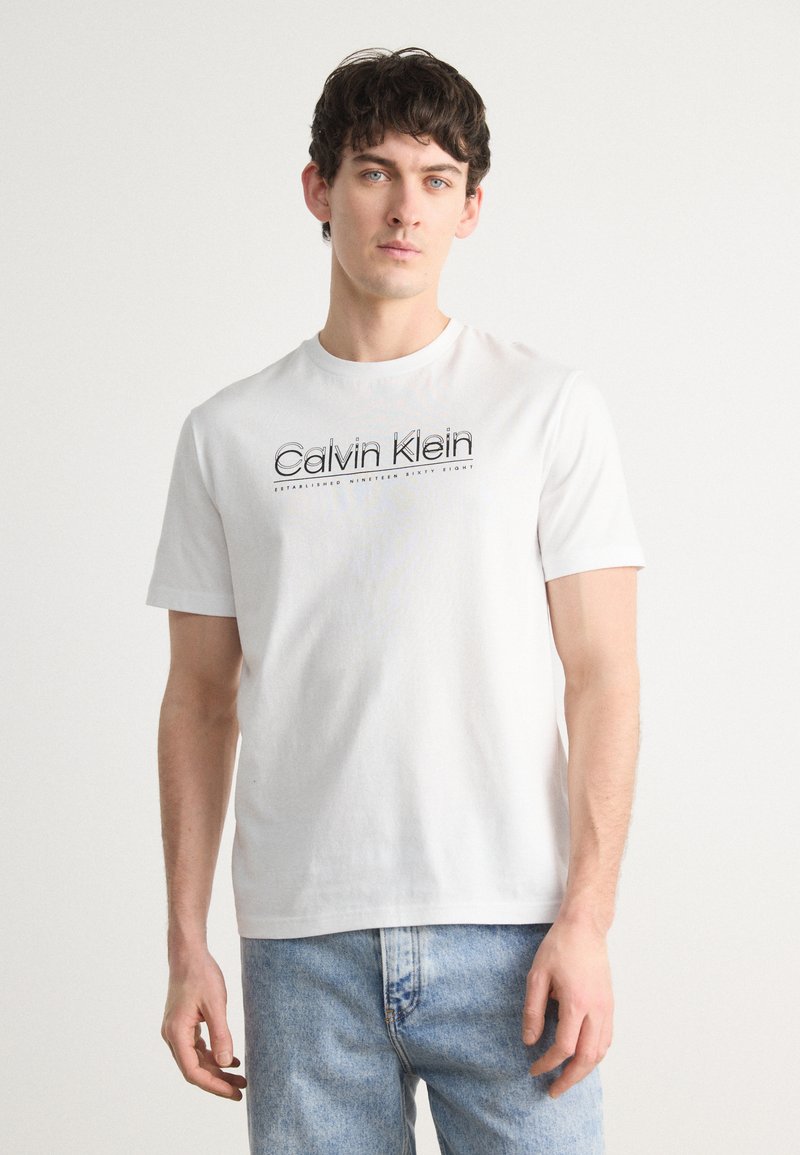 Balta trumpa rankovėmis marškinėliai pagaminti iš medvilnės, su juodai spaustu "Calvin Klein" logotipu ir tekstu. Dėvimi su šviesiai mėlynomis džinsinėmis kelnėmis.