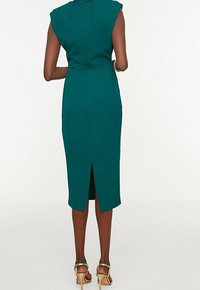 Robe ajustée turquoise avec détails aux épaules et une fente au dos, confectionnée dans un tissu lisse. Portée avec des sandales à talons dorés. Design minimaliste.