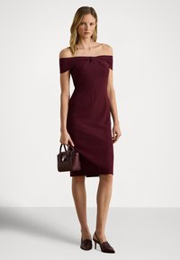 Bordové šaty s odhalenými ramenami, priliehavým strihom a uzlovým detailom vpredu, doplnené o malú zladenú kabelku a špicaté lodičky.