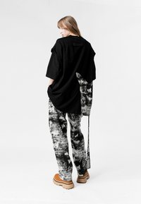 Joven con cabello largo, vistiendo ropa de estampado abstracto en blanco y negro de gran tamaño y zapatos de suela gruesa en color beige, de pie con las manos en los bolsillos.