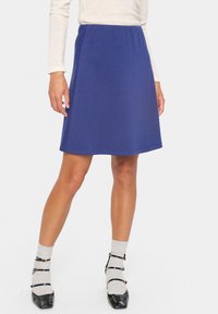 Jupe bleu mi-longue en A en tissu doux, avec une taille élastique. Associée à des escarpins noirs en vernis et des chaussettes grises.