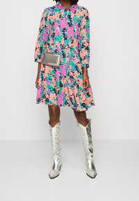 Donna che indossa un vestito floreale colorato, stivali metallici argento fino al ginocchio e una piccola borsa a tracolla argento.