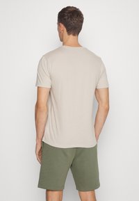 Camiseta beige de manga corta con escote redondo, combinada con pantalones cortos verdes. La tela parece suave y ligera, con una textura lisa.